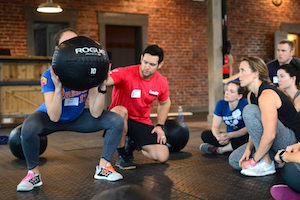 A small group of prospective CrossFit trainers and athletes observe a fellow aspiring trainer learn the fundamentals of the press with PVC as a Level 1 Course instructor coaches her.