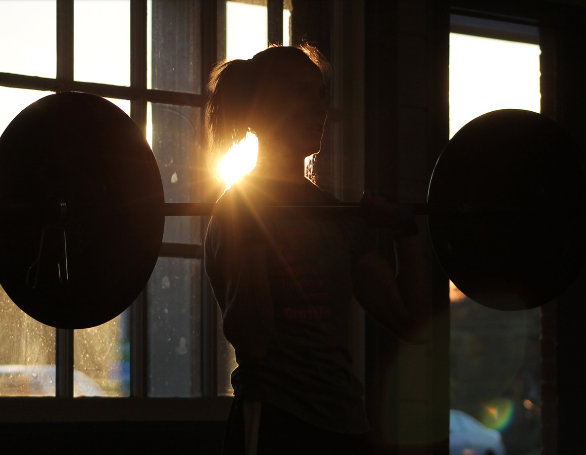 The silhouette of a woman holding a barbell, backlit by the sun