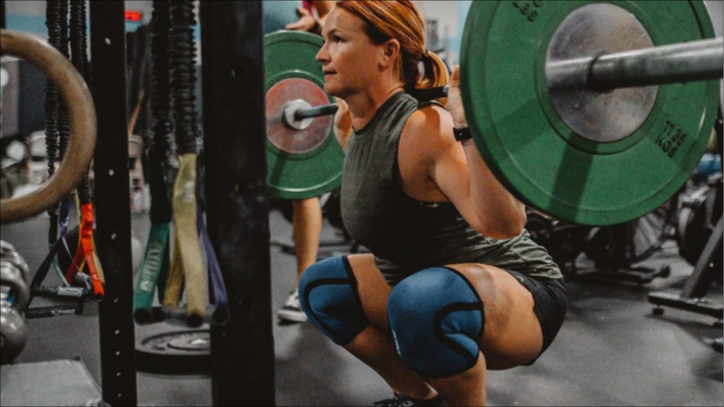A muscular athlete performs a single-arm dumbbell snatch, a common CrossFit exercise, holding the weight overhead in a low